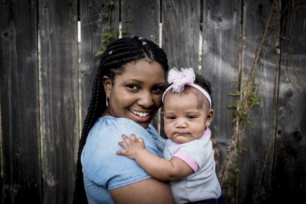 A woman with braided hair smiles while holding a baby wearing a pink headband. They are standing in front of a wooden fence with vines.
